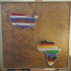 May include: A wooden wall art piece with string art depicting the outlines of the United States and Africa. The outlines are filled with the colors of the American and African flags. A red heart connects the two continents.
