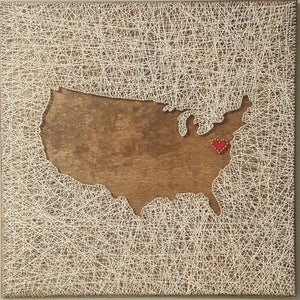 May include: A string art piece depicting the outline of the United States with a red heart marking a location in the eastern part of the country.