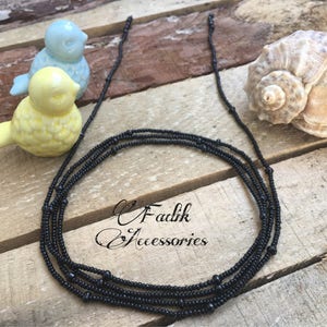 May include: A black beaded necklace with multiple strands. The necklace is made of small, round beads and is lying on a wooden surface.