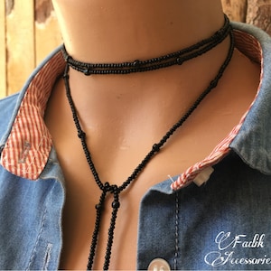 May include: A black beaded necklace with a long tassel. The necklace is layered and has a simple design. The necklace is worn by a person wearing a denim shirt with a red and white checked collar.
