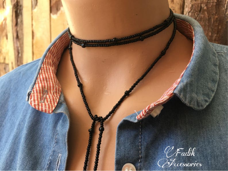 May include: A black beaded necklace with a long tassel. The necklace is layered and has a simple design. The necklace is worn by a person wearing a denim shirt with a red and white checked collar.