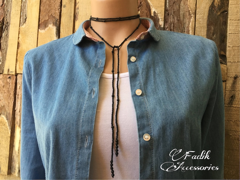 May include: A black beaded necklace with a long pendant. The necklace is being worn by a person wearing a denim shirt.