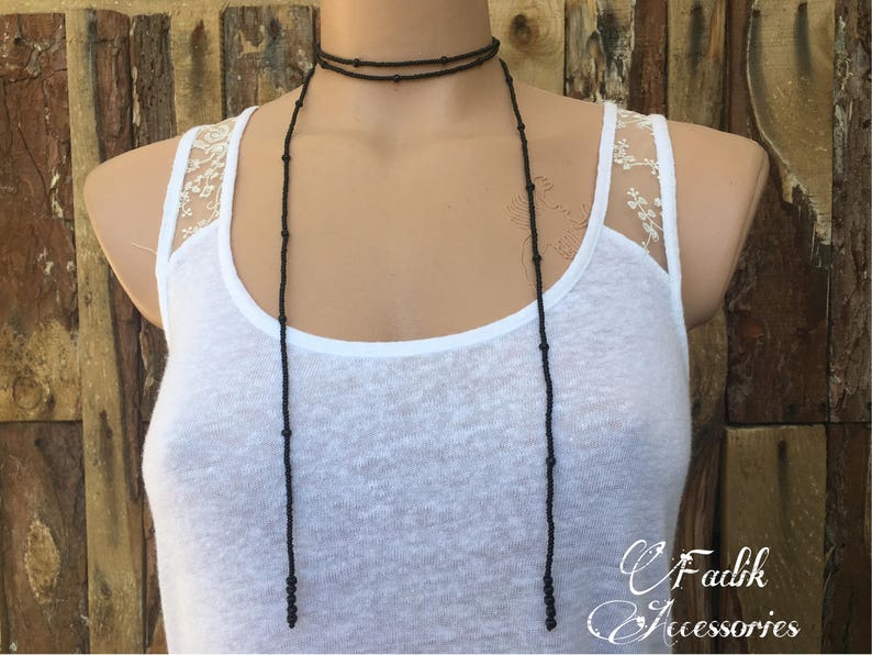 May include: A long black beaded necklace with two strands. The necklace is worn by a person wearing a white tank top with a lace overlay.