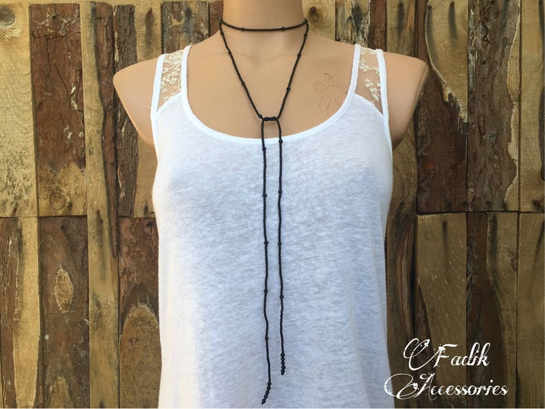 May include: A black beaded choker necklace with a long, dangling pendant. The necklace is worn by a person wearing a white tank top with lace trim.