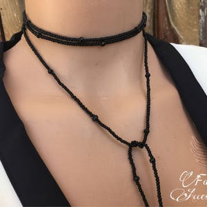 May include: A black beaded choker necklace with a long, dangling chain. The necklace is made of small, round beads and has a simple, elegant design.