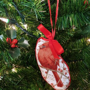 May include: Red cardinal bird Christmas ornament with a white background and red berries. The ornament is hanging on a green artificial Christmas tree.
