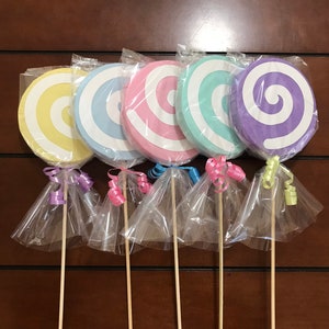May include: Five colorful, swirl-patterned lollipops in pastel shades of yellow, blue, pink, teal, and purple. Each lollipop is wrapped in clear cellophane and tied with a ribbon. The lollipops are attached to wooden sticks.