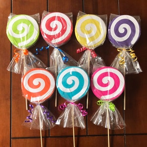May include: Eight large lollipop centerpieces with colorful swirls on a white background. The lollipops are on sticks and wrapped in clear cellophane with colorful ribbon.