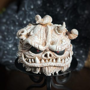May include: A white ceramic sculpture of a grotesque monster with red eyes and a toothy grin. The sculpture is sitting on a black metal stand.