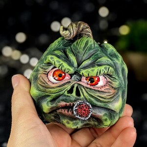 May include: A green pumpkin with a scary face, red eyes, and a cigar in its mouth. The pumpkin is hand-painted and has a detailed texture.