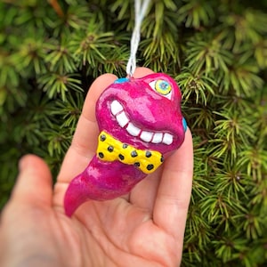 May include: A whimsical, handmade ornament in the shape of a smiling, magenta-colored creature with a yellow bow tie with black polka dots. The ornament has a white string for hanging and is set against a backdrop of green foliage.