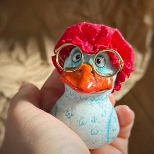 May include: A quirky ceramic duck figurine, hand-painted with a light blue body and an orange beak. It wears a red bonnet with white spots and gold-rimmed spectacles. The duck is held in a hand.