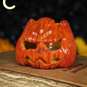 May include: Orange ceramic jack-o'-lantern with a carved face, lit from within. The pumpkin sits on a vintage book. The letter "C" is in the upper left corner. A Halloween decoration.