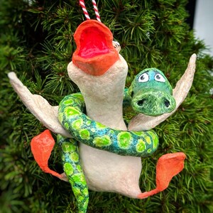May include: A whimsical handmade ornament featuring a white bird with an open orange beak and orange feet. A green and blue spotted snake wraps around the bird. The ornament is suspended by a red and white striped string, perfect for holiday decor.