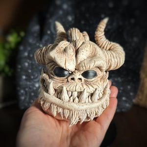 May include: A white ceramic sculpture of a pumpkin-like creature with horns, a wide grin, and black eyes. The sculpture is detailed with textured skin and sharp teeth.
