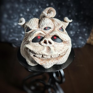 May include: A white ceramic pumpkin with a grotesque face, three eyes, and a toothy grin. The pumpkin is sitting on a black metal stand.