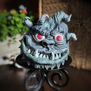 May include: A green and gray sculpted demon head with red eyes and sharp teeth. The head is sitting on a black metal stand.