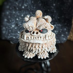 May include: A white ceramic monster figurine with red eyes and a toothy grin. The monster has a shaggy white fur-like texture and is standing on a black pedestal.