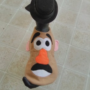 May include: A brown felt toy with a Mr. Potato Head face, wearing a black felt hat. The toy has a large orange nose, black eyebrows, and white eyes.