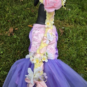 Pode incluir: Um vestido de princesa roxo e rosa com uma trança amarela e detalhes florais. O vestido apresenta um corpete rosa com um padrão xadrez e uma saia de tule roxa em camadas. A trança é adornada com flores e fitas, e uma grande rosa rosa está no topo.