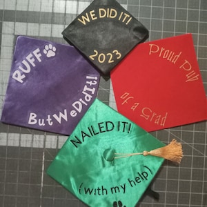 May include: Four graduation caps with different designs and colours. The caps are decorated with paw prints and text, including "RUFF", "But We Did It!", "We Did It! 2023", "Proud Pup of a Grad", and "NAILED IT! (with my help)".