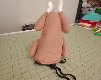 May include: A brown plush turkey with white felt wings and a black elastic strap. The turkey is sitting on a gray cutting mat.