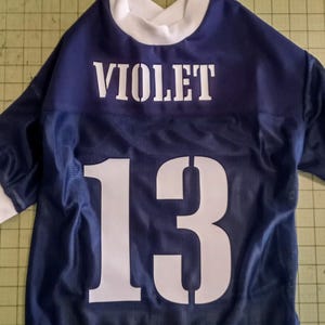 Pu&ograve; includere: Una maglia blu navy con tessuto a rete bianca. La maglia ha il nome "VIOLET" e il numero "13" stampato sul retro in bianco.