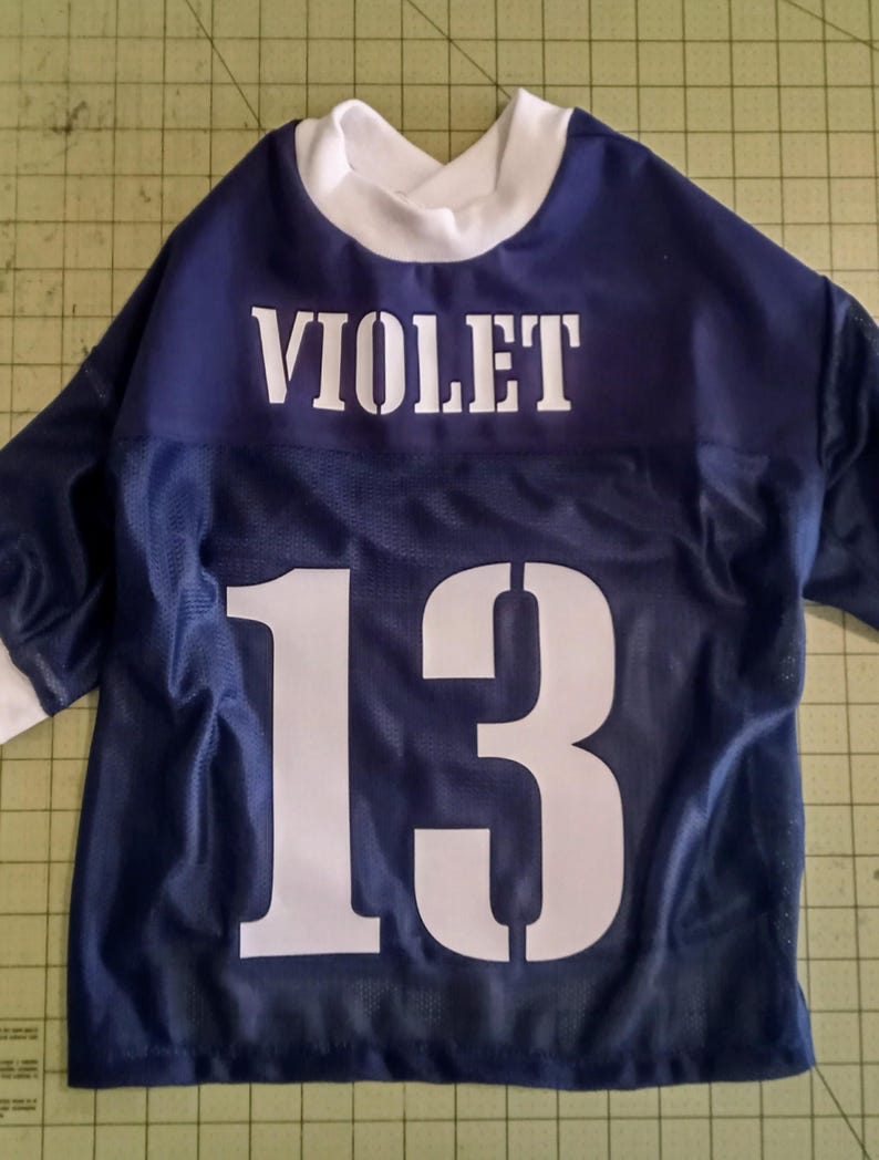 Pu&ograve; includere: Una maglia blu navy con tessuto a rete bianca. La maglia ha il nome "VIOLET" e il numero "13" stampato sul retro in bianco.