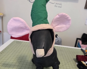 May include: A black dog costume with a green elf hat, pink ears, and a beige muzzle. The costume is designed to resemble a mouse or a similar creature. The dog is standing on a green cutting mat.