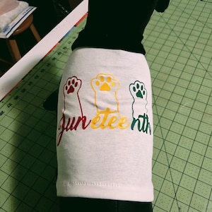 May include: White t-shirt with the word "Juneteenth" in red, yellow, and green script, accompanied by paw print designs in matching colors. The shirt is displayed on a black surface.