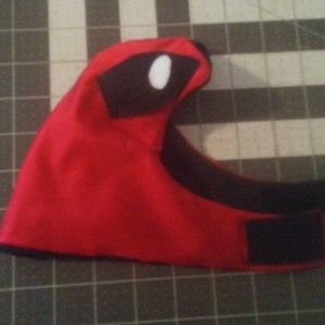 May include: Red and black fabric mask with a white eye detail. The mask has a black strap with velcro closures.