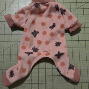 May include: A pink pet onesie featuring a pattern of orange polka dots and dark grey insect motifs. The garment has a high collar, short sleeves, and covers the legs. The cuffs and collar are a darker shade of pink.