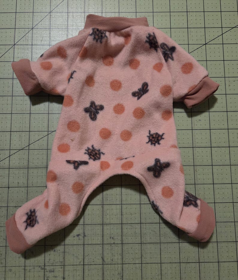 May include: A pink pet onesie featuring a pattern of orange polka dots and dark grey insect motifs. The garment has a high collar, short sleeves, and covers the legs. The cuffs and collar are a darker shade of pink.