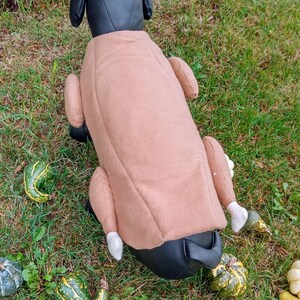 以下が含まれることがあります： ローストターキーの形をした茶色のぬいぐるみ製の犬用コスチューム。コスチュームは、ターキーの脚と羽根に白いアクセントが付いています。犬用コスチュームは、緑の芝生の上に置かれ、周りに小さなカボチャが散らばっています。