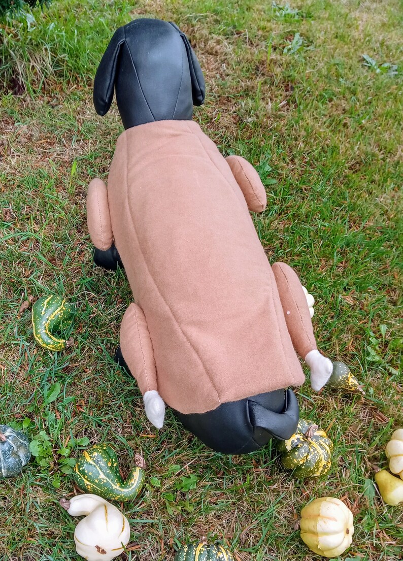 以下が含まれることがあります： ローストターキーの形をした茶色のぬいぐるみ製の犬用コスチューム。コスチュームは、ターキーの脚と羽根に白いアクセントが付いています。犬用コスチュームは、緑の芝生の上に置かれ、周りに小さなカボチャが散らばっています。