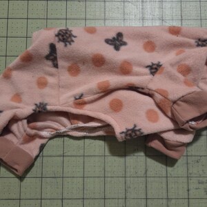 May include: Pink fleece pet pyjamas with a pattern of orange polka dots and dark grey bee and anchor designs. The pyjamas have brown cuffs and trim. The garment is folded on a green grid surface.