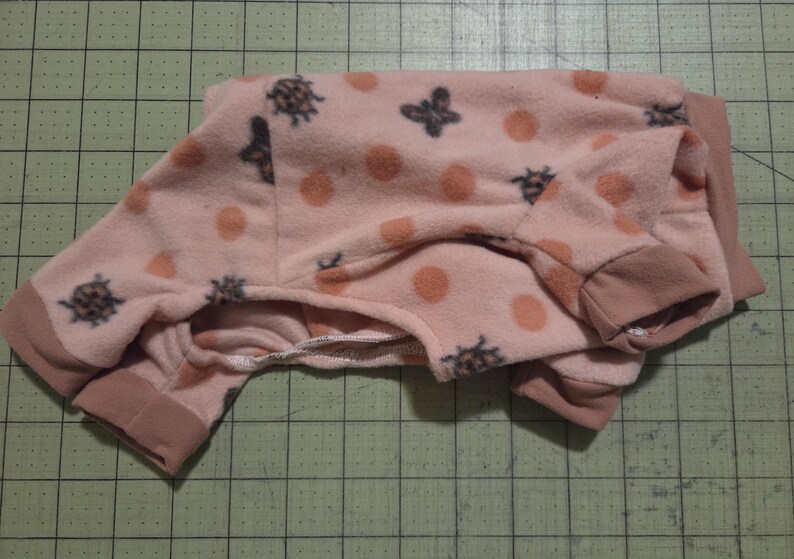 May include: Pink fleece pet pyjamas with a pattern of orange polka dots and dark grey bee and anchor designs. The pyjamas have brown cuffs and trim. The garment is folded on a green grid surface.