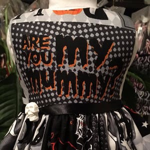 May include: A black and white dress with orange and red accents. The dress has a skull charm on the belt and the text "Are you my mummy?" on the bodice. The skirt has a Halloween theme with ghosts, bats, and pumpkins.