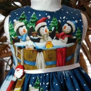 Puede incluir: Un vestido azul sin mangas con temática de pingüinos y Navidad. El vestido tiene un cinturón de cinta blanca y un botón rojo de Papá Noel.
