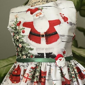May include: A white sleeveless dress with a green sash and a red and white Santa Claus print. The dress has a full skirt and is decorated with pine trees, presents, and other Christmas-themed imagery.