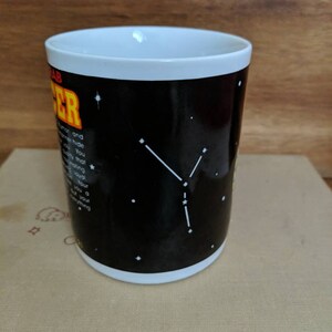 May include: A white-rimmed coffee mug with a black background featuring the Cancer zodiac sign. The mug has white constellations and text describing the Cancer personality. The mug is sitting on a stack of books.
