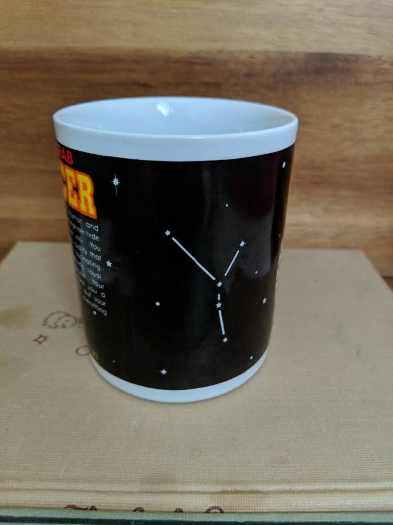 May include: A white-rimmed coffee mug with a black background featuring the Cancer zodiac sign. The mug has white constellations and text describing the Cancer personality. The mug is sitting on a stack of books.