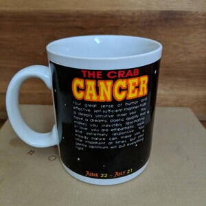 May include: White ceramic mug with a black background featuring the text "THE CRAB CANCER" in yellow and red. The mug includes a description of the Cancer zodiac sign and the dates June 22 - July 21.