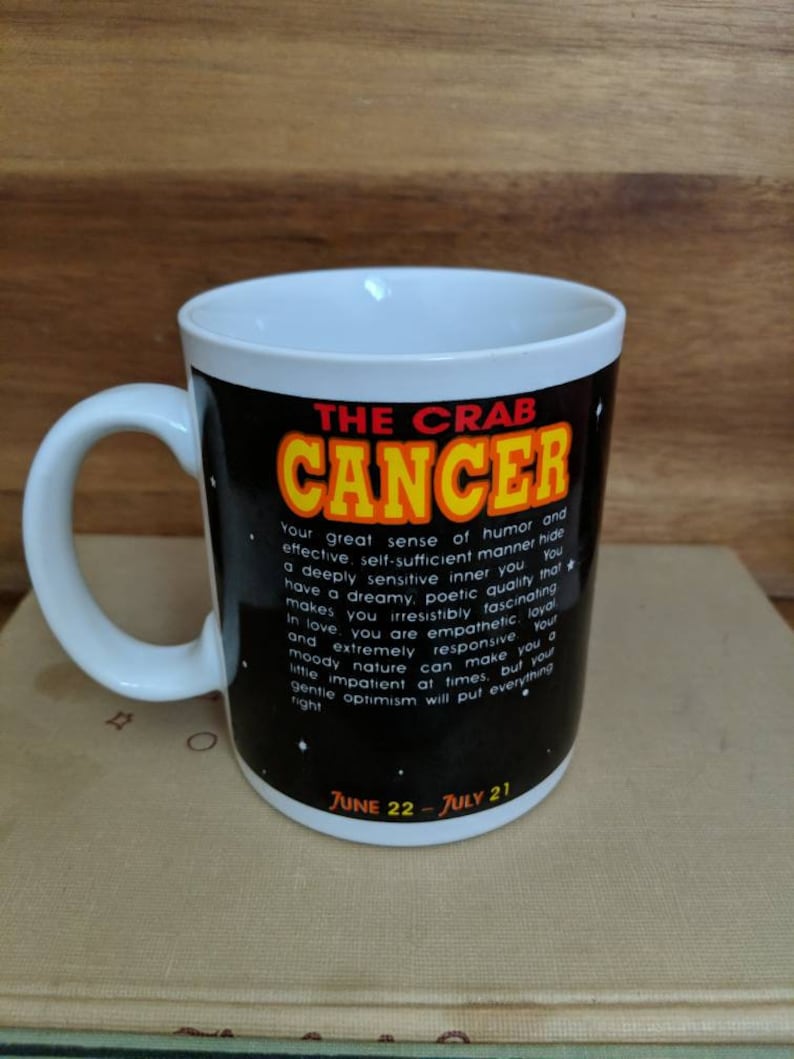 May include: White ceramic mug with a black background featuring the text "THE CRAB CANCER" in yellow and red. The mug includes a description of the Cancer zodiac sign and the dates June 22 - July 21.