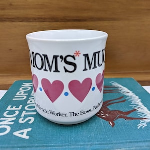 May include: White ceramic mug with the words "MOM'S MUG" in black, accented with pink hearts and blue dots. Additional text reads "Miracle Worker, The Boss, Pushover". A charming gift for Mother's Day or any occasion.