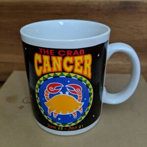May include: White ceramic mug with a black background featuring the Cancer zodiac sign. The mug has a crab illustration in yellow and red, with the words "THE CRAB CANCER" in yellow and orange. The dates "JUNE 22 - JULY 21" are also printed on the mug.