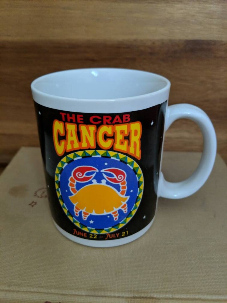 May include: White ceramic mug with a black background featuring the Cancer zodiac sign. The mug has a crab illustration in yellow and red, with the words "THE CRAB CANCER" in yellow and orange. The dates "JUNE 22 - JULY 21" are also printed on the mug.