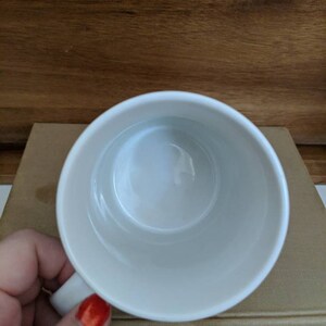 May include: A white ceramic mug with a handle, viewed from above. The interior of the mug is smooth and clean. The mug is resting on a stack of books with a wooden background.