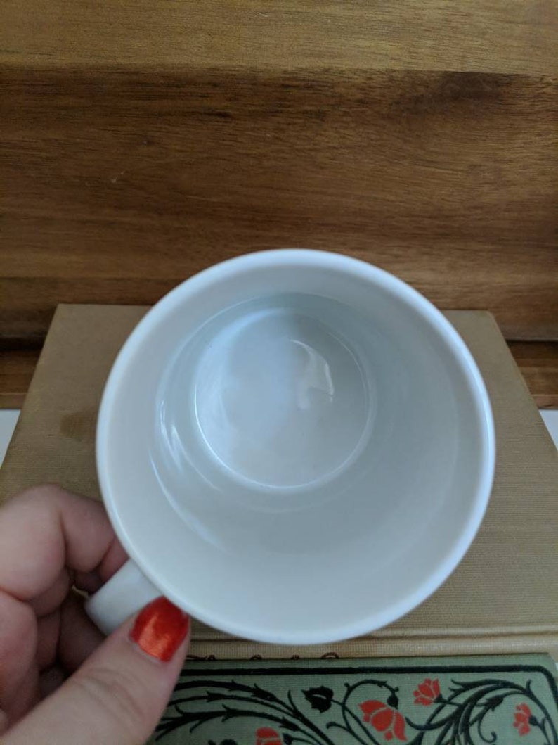 May include: A white ceramic mug with a handle, viewed from above. The interior of the mug is smooth and clean. The mug is resting on a stack of books with a wooden background.