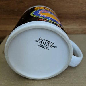 May include: White ceramic mug with a black upper band featuring the word "CAMEL" and the dates "JUNE 22 - JULY". The bottom of the mug has the text "PAPEL FREELANCE MADE IN INDONESIA".